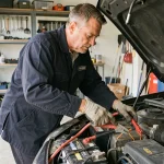 Homme en veste de mécanicien connectant des pinces de démarrage sur une batterie de voiture dans un garage résidentiel